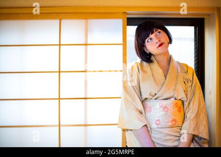 Maison japonaise traditionnelle avec femme en costume kimono robe regardant vers le haut avec de courts cheveux noirs sur le rebord de fenêtre de papier glissant en bois Banque D'Images