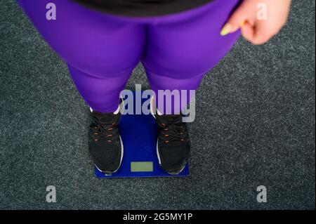 La femme grasse est pesée dans la salle de gym. Vue de dessus sur les jambes des femmes dans les baskets sur les balances électroniques. Obésité. Banque D'Images