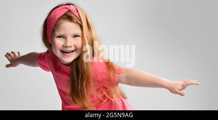 Riant redhead enfant fille dans les supports de t-shirt rose tenant les mains écartent, jouant oiseau, avion. Bannière Banque D'Images