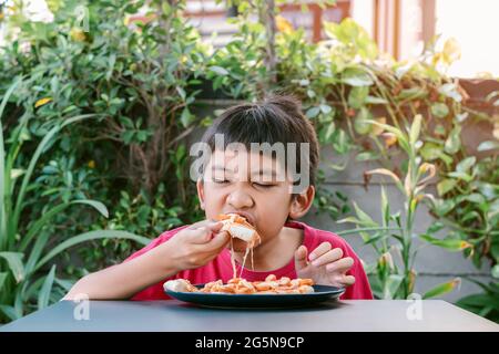 Petit garçon asiatique en chemise rouge manger une pizza délicieusement et heureusement. Banque D'Images