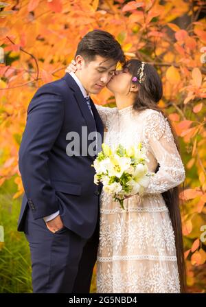 Couple biracial nouvellement marié embrassant à l'extérieur par des feuilles d'automne orange vif Banque D'Images