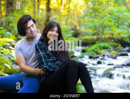 Jeune couple biracial assis le long d'un ruisseau rocheux dans des bois verts embrassant Banque D'Images