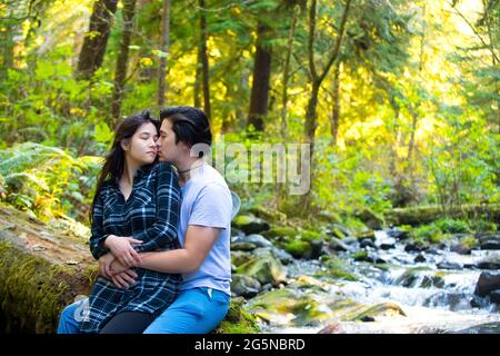 Jeune couple biracial assis le long d'un ruisseau rocheux dans des bois verts embrassant Banque D'Images