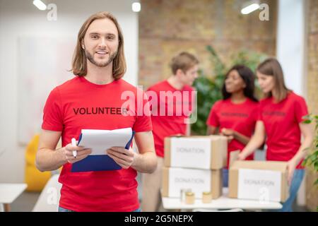 Guy dans un t-shirt de bénévole avec un document et des collègues derrière Banque D'Images