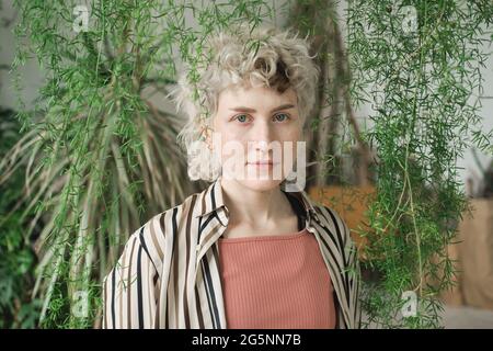 Portrait d'une jeune femme aux cheveux blonds et bouclés regardant un appareil photo debout parmi des plantes vertes Banque D'Images