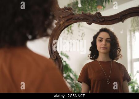 Jeune femme aux cheveux bouclés debout devant le miroir et regardant elle-même Banque D'Images