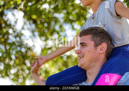 Frère défoqué de la sœur de circonscription à l'arrière. Portrait d'une fille heureuse sur les épaules de l'homme, porcgyback. Petite mouche. Famille jouant à l'extérieur. Arrière-plan de l'arbre vert. Banque D'Images