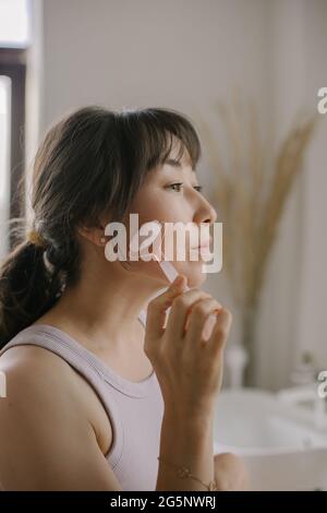 Vue latérale de la belle femme asiatique sans maquillage faire un massage du visage avec rouleau de quartz rose pendant la routine quotidienne de beauté matin, debout dans la salle de bains Banque D'Images