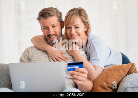 Achats en ligne à domicile. Beau homme tenant une carte bancaire dans sa main tandis que sa femme s'assoit à côté de lui et en utilisant un ordinateur portable tout en faisant des achats en ligne. Banque D'Images