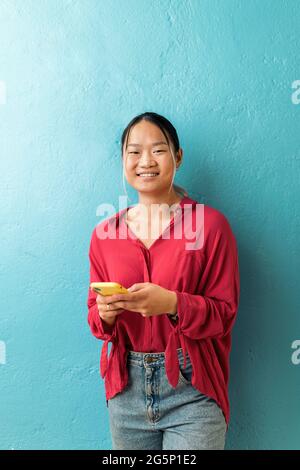 Une employée de bureau asiatique souriante tient son téléphone portable et regarde l'appareil photo sur fond bleu. Banque D'Images