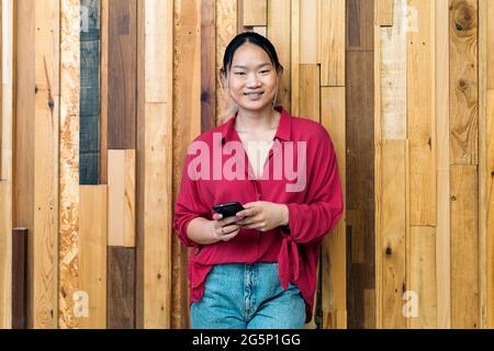 Une employée de bureau asiatique souriante tient son téléphone portable et regarde l'appareil photo sur fond de bois. Banque D'Images