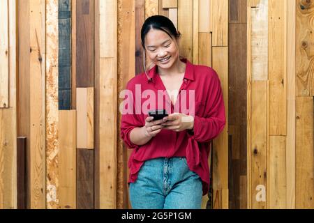 Une employée de bureau asiatique souriante tient son téléphone portable et tape sur un fond en bois. Banque D'Images