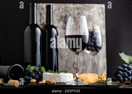 Vin rouge en verre classique avec composition de bouteilles de vin. Menu élégant du bar à vins avec en-cas au fromage sur fond sombre et moody. Vin rouge vintage sur fond noir Banque D'Images