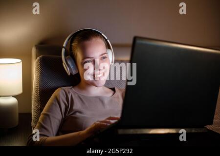 Une jeune femme heureuse dans un casque qui travaille ou étudie la nuit sur un canapé à l'aide d'un ordinateur portable. Adolescente se reposant studing regarder un film sur un ordinateur portable. internet Banque D'Images