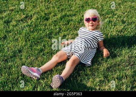 Petite fille heureuse en lunettes de soleil et une robe rayée se trouve sur l'herbe et sourit. Fille de mode posant en été à l'extérieur. Banque D'Images