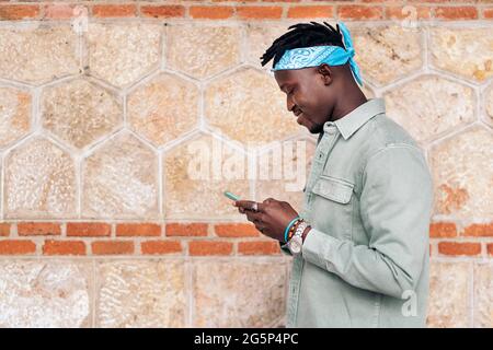 Jeune et élégant homme africain utilisant son téléphone portable contre le mur brun. Banque D'Images