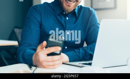Petit sans visage souriant jeune homme dans une tenue décontractée regardant l'écran de smartphone sur le lieu de travail Banque D'Images
