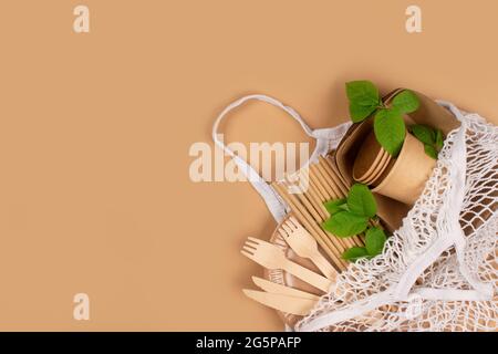 Tasses, assiettes et récipients en papier Kraft avec couverts en bois dans un sac en filet de coton blanc sur fond marron avec espace pour les copies. Repas à emporter dans la rue Banque D'Images