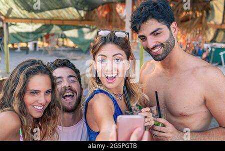 Rassemblement de jeunes en vacances souriant prenant un selfie sous un parapluie sur la plage en été Banque D'Images