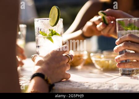 Mains tenant un cocktail de longue boisson alcoolisée frais sur une table. Rassemblement d'amis dans un pub en plein air en été. Banque D'Images