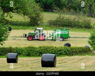 Production de foin ou d'ensilage (agriculteur travaillant dans un tracteur agricole sur une ramasseuse-presse à balles pour le travail dans les champs ruraux, collecte de l'herbe sèche et balles rondes enveloppées - Yorkshire England, Royaume-Uni. Banque D'Images