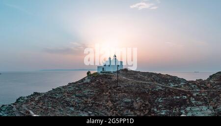 Phare et église d'Agios Nikolaos sur un cap rocheux, drone, vue aérienne. Coucher de soleil sur l'île de Kea Tzia. Grèce, Cyclades. Mer Égée rippée. Été v Banque D'Images