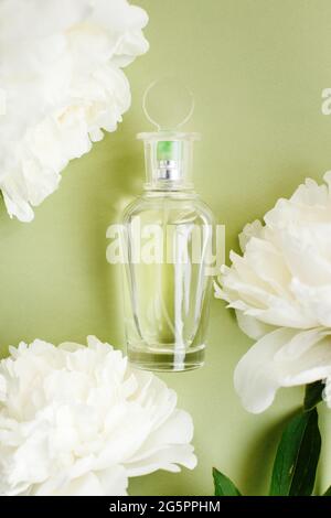 Bouteille en verre transparent avec parfum parmi les fleurs de pivoine blanc frais dans une couleur pastel vert olive, vue du dessus Banque D'Images