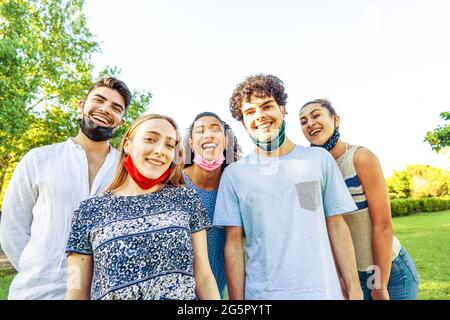 Heureux groupe insouciant d'amis multiraciaux posant dans un parc pour un portrait portant un masque de protection abaissé pour montrer leurs beaux sourires. Co Banque D'Images