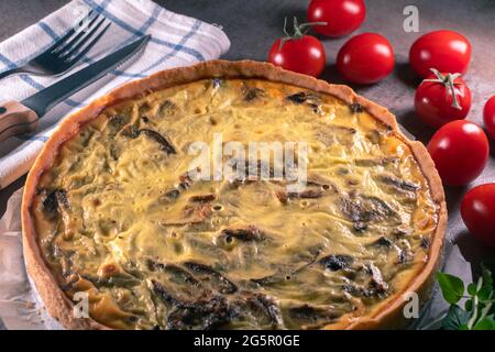 Gros plan d'une quiche jaune fraîchement cuite avec des champignons encore sur un papier de cuisson, assis sur une surface de table de cuisine sombre avec couteau, fourchette, tissu, toma Banque D'Images