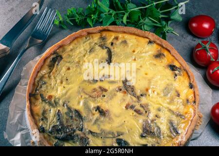 Gros plan d'une quiche jaune fraîchement cuite avec des champignons encore sur un papier de cuisson, assis sur une table de cuisine sombre avec couverts, t Banque D'Images