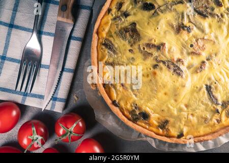 Vue de dessus d'une quiche jaune fraîchement cuite avec des champignons encore sur un papier de cuisson, assis sur une table de cuisine sombre avec couverts, tomates, Banque D'Images