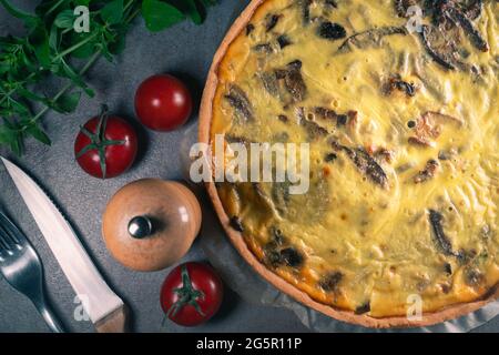 Vue de dessus d'une quiche jaune fraîchement cuite avec des champignons encore sur un papier de cuisson, assis sur une table de cuisine sombre avec couverts, tomates, Banque D'Images