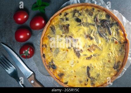 Vue de dessus d'une quiche jaune fraîchement cuite avec des champignons encore sur un papier de cuisson, assis sur une table de cuisine sombre avec couverts, tomates, Banque D'Images