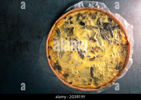 Vue de dessus d'une quiche jaune fraîchement cuite avec des champignons encore sur un papier de cuisson, assis sur une table de cuisine sombre Banque D'Images