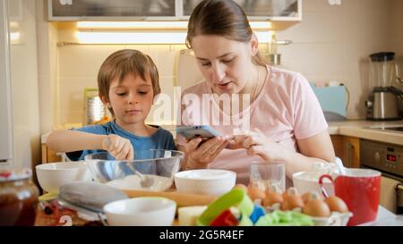 Joyeux garçon souriant nourrissant sa mère à l'aide d'une cuillère tout en cuisinant une tarte ou un gâteau à la maison. Les enfants cuisent avec leurs parents, le petit chef, la famille ayant du temps ensemble Banque D'Images