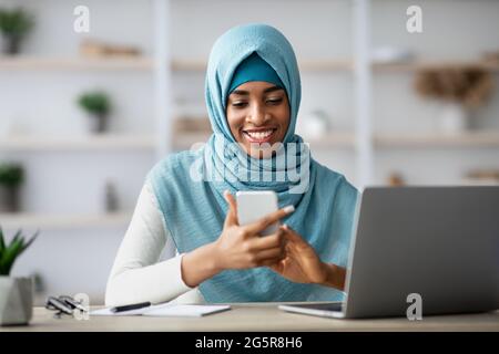 Femme d'affaires noire dans hijab utilisant le téléphone portable tout en étant assis au bureau avec ordinateur portable Banque D'Images