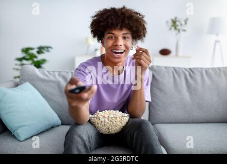 Divertissements à la maison. Adolescent afro-américain enthousiaste avec une télécommande pour regarder un film et manger du pop-corn à l'intérieur Banque D'Images