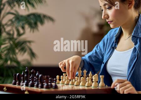 Jeune femme regardant les pièces d'échecs sur le plateau et se déplaçant tout en jouant au jeu de plateau d'échecs à la maison seule Banque D'Images