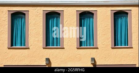 Quatre fenêtres à ombre avec rideaux bleu sarcelle coulant sur un bâtiment en stuc de couleur Terra Cotta. Banque D'Images