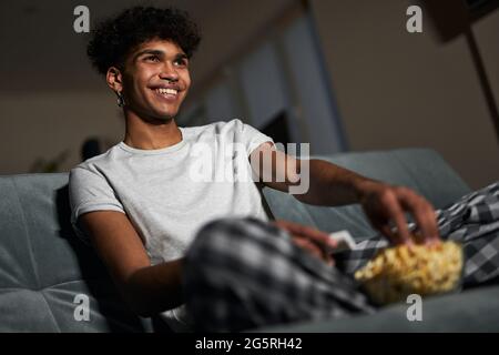 Un jeune garçon joyeux portant un pyjama qui prend du pop-corn dans un bol tout en regardant la comédie, assis sur un canapé à la maison Banque D'Images