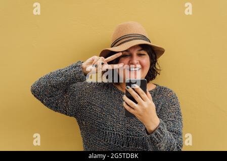 Une femme a accueilli par son téléphone cellulaire. Une fille qui a un appel vidéo sur son smartphone, porte un chapeau et est sur un mur de couleur moutarde Banque D'Images