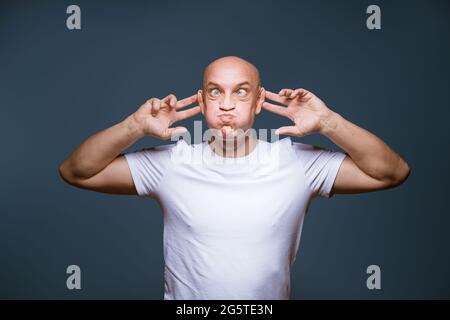 Homme à tête chauve sur fond bleu avec des expressions du visage excitées et des mains levées à sa tête un homme caucasien grimaquant dans un t-shirt blanc Banque D'Images