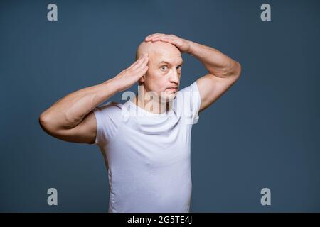 Drôle de chauve homme posant sur un fond bleu avec des expressions du visage excitées et des mains levées à sa tête Caucasien gars grimacing dans le t-shirt blanc Banque D'Images