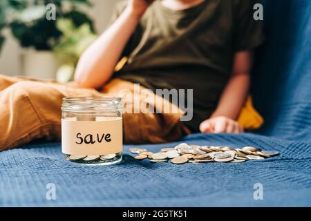 Gros plan sur les mains de petit enfant garçon qui attrapant et mettant des pièces de monnaie empilées dans un pot en verre avec une étiquette de sauvegarde. Don, économie d'argent, charité, famille Banque D'Images