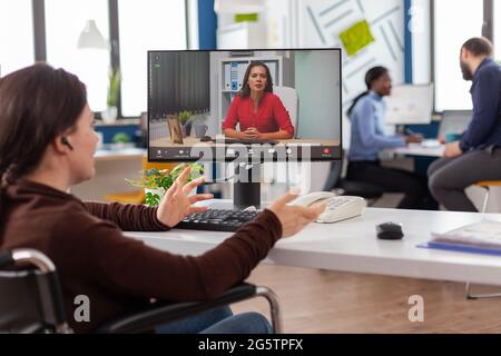 Invalide invalide femme d'affaires immobilisée parlant de rapport de vente dans la visioconférence travaillant des heures supplémentaires dans le bureau de démarrage. Équipe utilisant la communication sans fil en ligne discuter sur webcam pendant la réunion de groupe. Banque D'Images