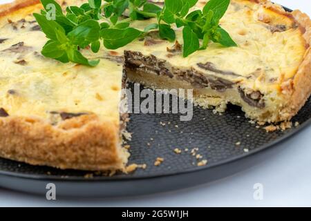 Gros plan grand studio de gâteau salé français jaune fraîchement cuit, ou quiche, avec des champignons et de la menthe verte, sur une assiette design noire, isolée sur le whi Banque D'Images