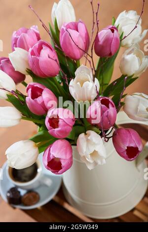 Un bouquet de tulipes hollandaises blanches et roses avec du bouleau rose dans un vase en pot blanc sur une table basse avec une petite tasse de café dans un cadre familial Banque D'Images