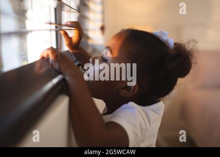 Curieuse fille afro-américaine debout et peering à travers les stores de fenêtre sur un jour ensoleillé Banque D'Images