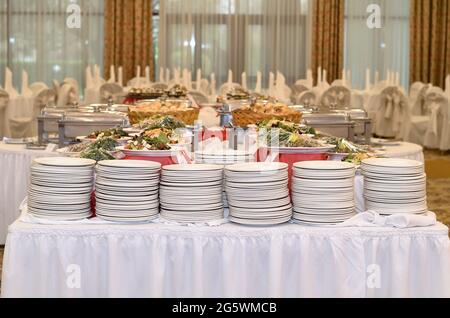 Table de restauration avec une nappe blanche et six piles où il y a beaucoup de plats blancs et salades préparées dans une grande salle de banquet avec fenêtres Banque D'Images