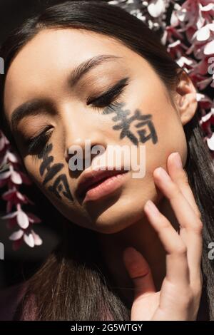 Portrait d'une femme asiatique avec hiéroglyphes sur le visage et kanzashi isolé sur le noir Banque D'Images